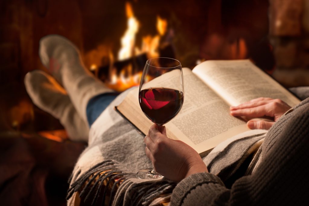 Lady sitting in front of fire with book and glass of wine with feet up