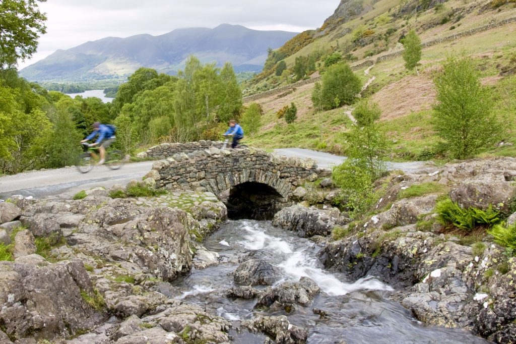 Two cyclists going over bridge that straddles stream with mountains in back ground