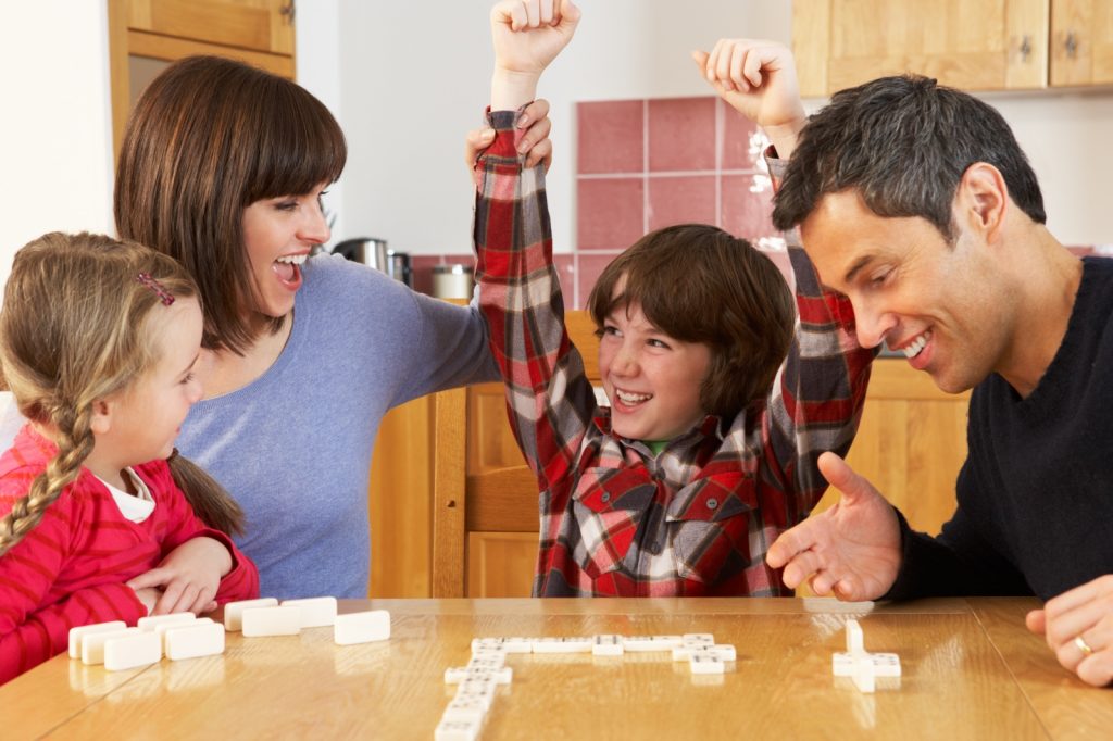 Family sitting around table playing scrabble with boy throwing arms in air celebrating victory