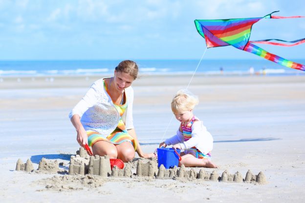 Mother and small child sitting on a sandy beach building sand castles with colourful kite flying above