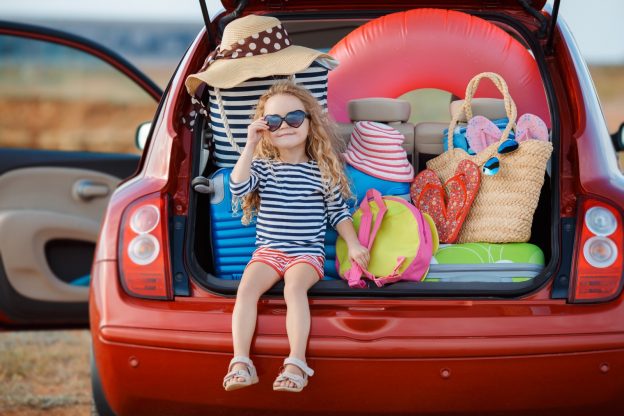Little girl with heart shaped glasses on sitting in the boot of a car stacked with cases, sun hats and beach gear