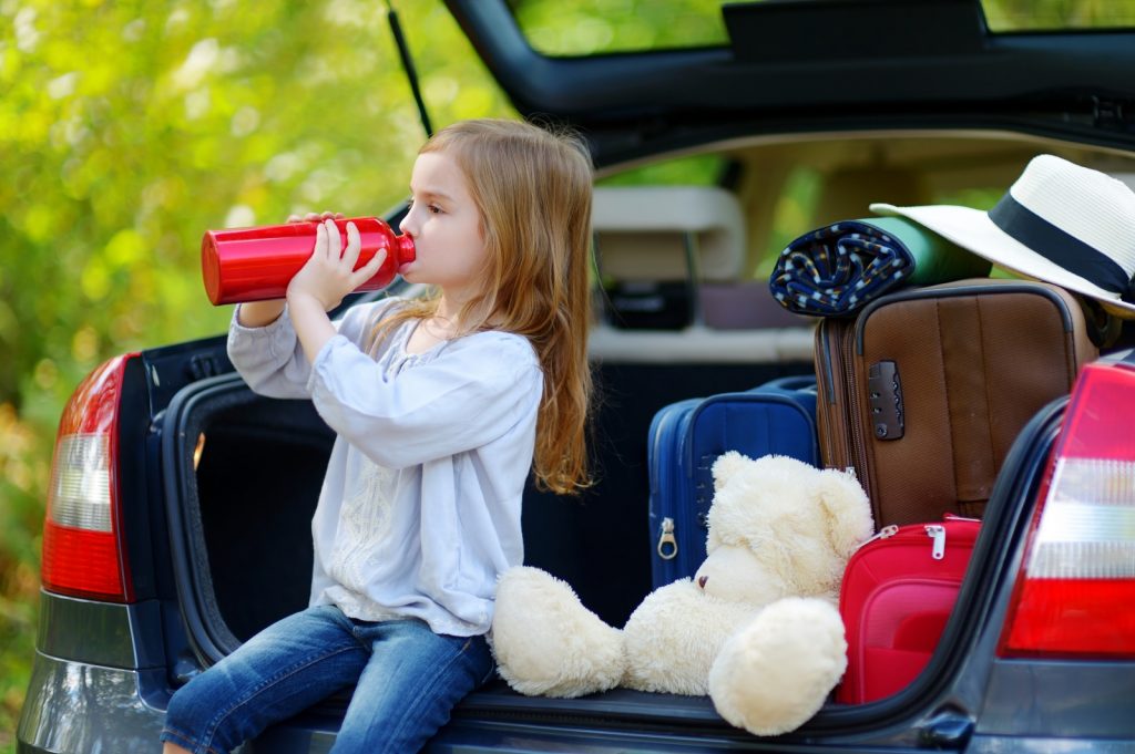 Little girl sitting in the boot of a car drinking out of a bottle with teddy and suitcases