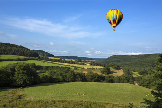 Colourful hot air balloon in bright blue sky over sheep in green fields and trees